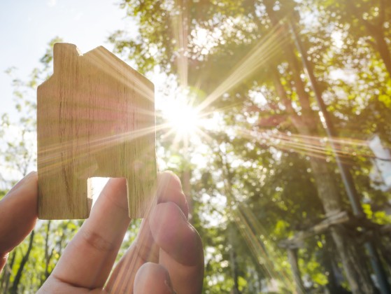 eine Hand hält eine kleine Figur von einem Haus ins Sonnenlicht, im Hintergrund Bäume